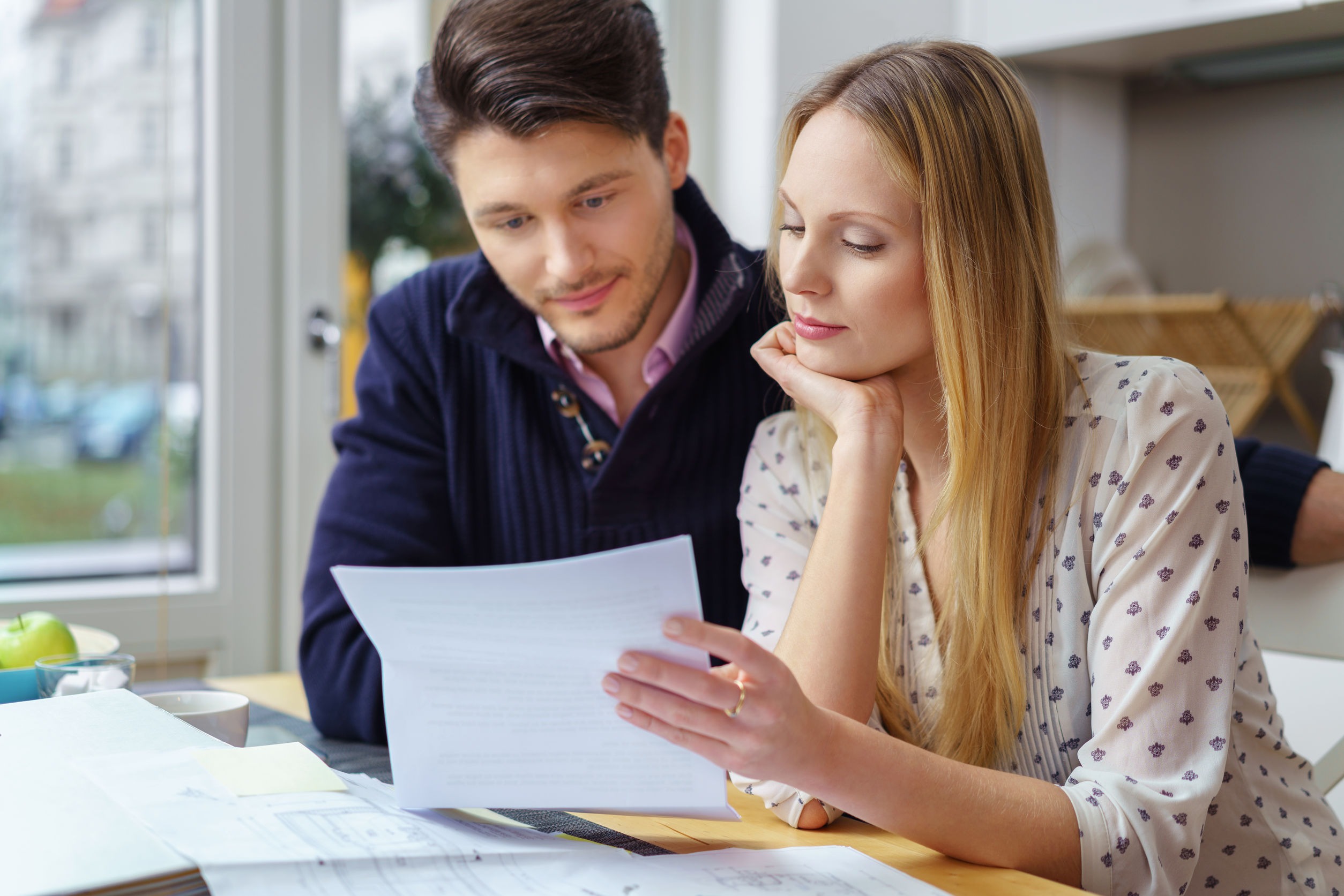 Handsome young man with mustache and beautiful woman with long hair at table looking at documents in kitchen next to window